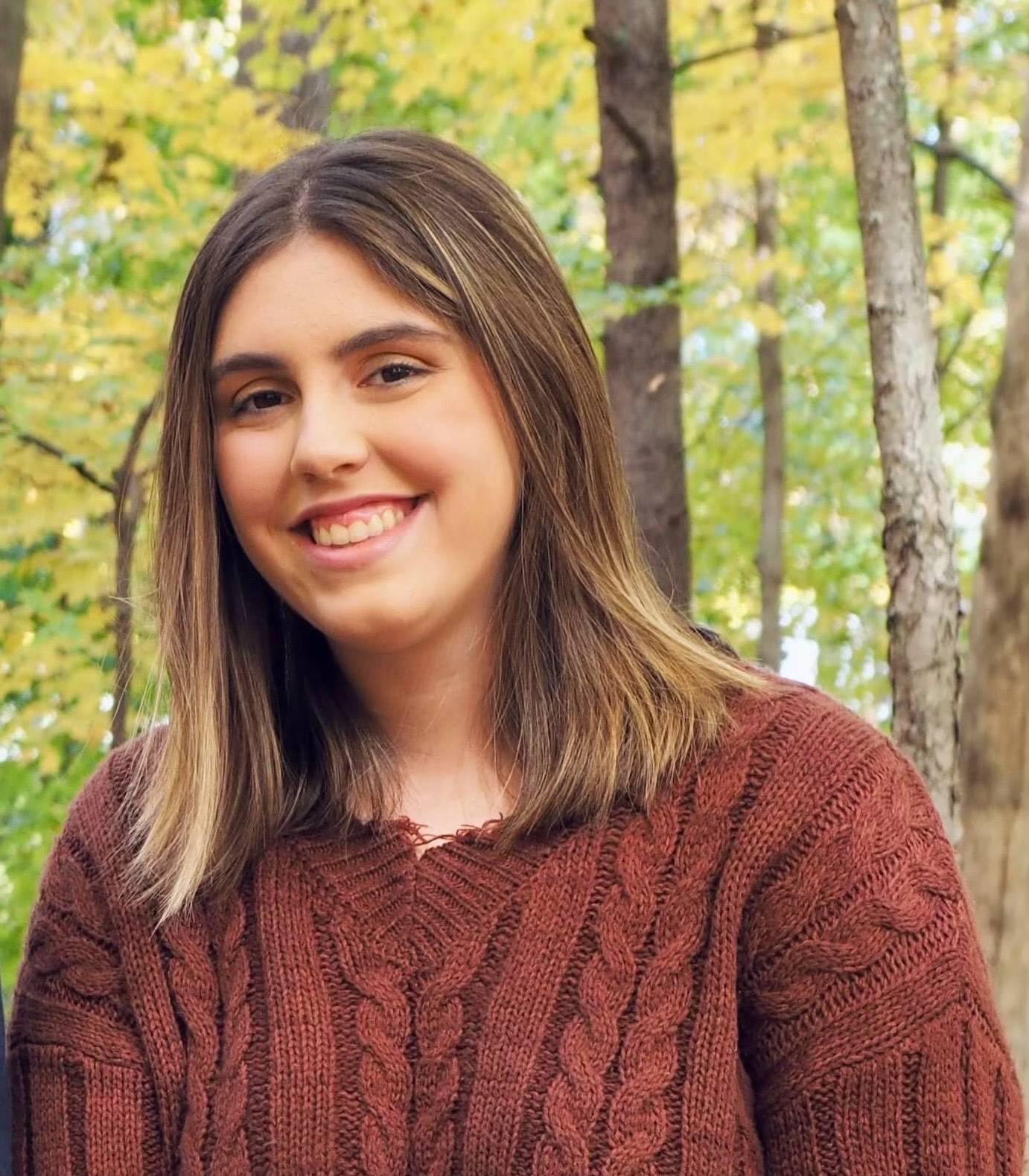 Girl with brown hair and burgundy knitted sweater smiling at the camera with trees in the background
